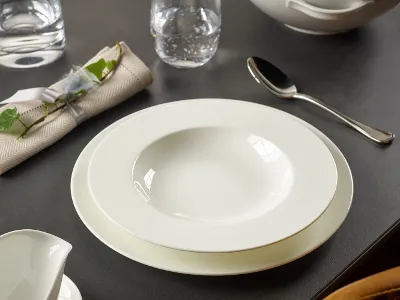 The table setting consists of a white Anmut bowl from Villeroy & Boch on a plate, a spoon, a folded napkin decorated with greenery, and a glass of water, all arranged on a dark surface.