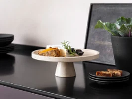 A selection of cheese on a Travertine serving platter with a foot, and dark fruits, next to black Manufacture tableware by Villeroy & Boch on a dark worktop.