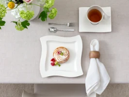 Dessert auf einem quadratischen NewWave Teller mit Himbeeren, Serviette, Löffel, Gabel und einer Tasse Tee auf einem Tisch mit heller Tischdecke und Blumenschmuck.