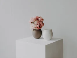A brown vase with pink flowers and a white ceramic vase stand on a white cube against a plain light-coloured background.