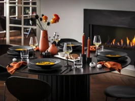 A modern dining table for five with black Manufacture dinnerware, orange napkins, and autumnal decorations, including vases of flowers and lit candles, next to a burning fireplace.