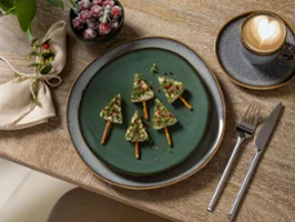 Ein Villeroy & Boch Lave Vert Dessertteller, dekoriert mit essbaren grünen Weihnachtsbäumen, platziert auf einem beigen Lave Teller mit passender Cappuccinotasse. Umgeben von festlicher Weihnachtsdekoration, die eine warme und rustikale Atmosphäre schafft.