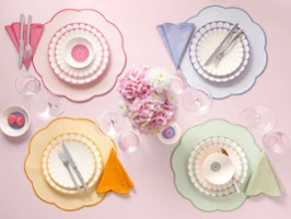 Four placemats with colorful plates and bowls from the Fleur collection, colorful napkins, and cutlery are arranged around a pink floral decoration.