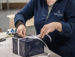 A person ties a white ribbon around a dark blue gift box on a table and prepares to wrap it.