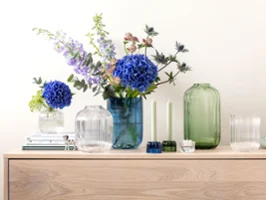 A sideboard with Fleur vases from Villeroy & Boch, decorated with blue and purple flowers, next to two green candles and stacked books.