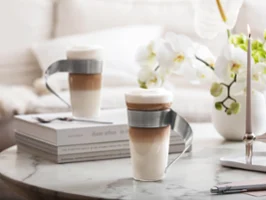 NewWave - Disfruta del café con elegancia Dos lattes en capas en modernas tazas de cristal NewWave con asas de metal curvadas se encuentran sobre una mesa de mármol con libros, flores y velas.