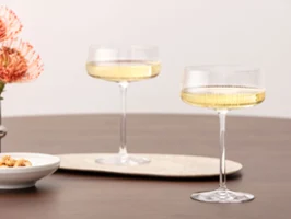 Two Afina glasses filled with sparkling wine on a wooden table, next to flowers and a small bowl.