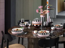 Ein runder Marmortisch mit vier Gedecken, gestreiftem Memphis Geschirr, Weingläsern und einer Vase mit rosa Blumen.