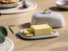 A rectangular block of butter with two slices lies on a ceramic Perlemor butter dish with a matching lid on a wooden table.