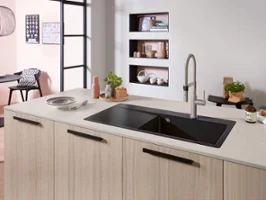 Una cucina moderna con un lavello nero su un'isola in legno chiaro, un rubinetto, una pianta e vari oggetti.