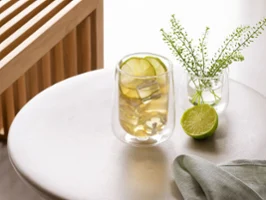 An Artesano glass with a chilled drink and slices of lime on a round table, next to it a vase with green plants and a halved lime.