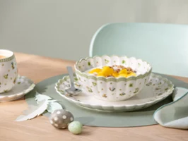 A decorative Easter Delight bowl with fruit and yogurt stands on a plate with a spoon next to a cup and saucer on a table.