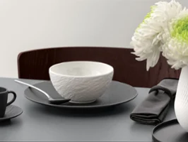 A formal table setting with a white bowl on a black plate and silver cutlery on a grey table.