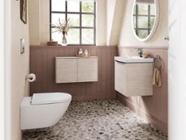 Une salle de bains avec une fenêtre, un sol en galets, un lavabo mural et des toilettes. Au-dessus du lavabo se trouve un miroir rond, les murs inférieurs sont recouverts de boiseries et des tableaux encadrés sont accrochés sur les murs supérieurs.