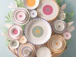 A collection of decorative plates from the Fleur collection with colourful patterns arranged on a wall and surrounded by paper leaf decorations.