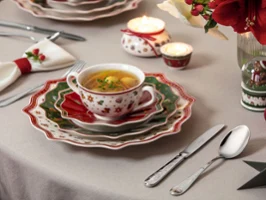 A festively set table with a soup cup and various plates A festively set table with a cup of soup, layered festive plates, cutlery, candles, and holiday decorations.