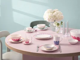 A dining table with pastel-coloured crockery, consisting of plates, bowls, cups, saucers and cutlery. A vase with white flowers and two candles stand on a light pink tablecloth.