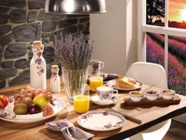 Ein Frühstücksgedeck mit dem Villeroy & Boch Artesano Provençal Lavendel-Geschirr, geschmückt mit Obst, einem Croissant, Müsli und Orangensaft. Eine Vase mit Lavendel steht elegant in der Mitte des Tisches. Das Fenster bietet einen malerischen Blick auf einen Sonnenuntergang über einem Feld aus lila Blumen.