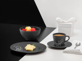 Czarna zastawa stołowa z kolekcji Manufacture Mickey obok białego pudełka prezentowego Czarna zastawa stołowa z kolekcji Manufacture Mickey z owocami, kanapką z masłem i kawą, obok białe pudełko prezentowe, serwetka i łyżka na podzielonym tle.