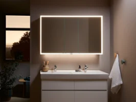 Une salle de bains moderne avec un grand miroir lumineux au-dessus d’un lavabo blanc et d’un meuble sous plan. Une serviette est accrochée au mur et divers articles de toilette se trouvent sur la tablette.