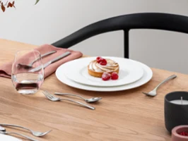 Ein mit Himbeeren garniertes Dessert steht auf einem Royal Teller mit Besteck auf einem Holztisch. Daneben stehen ein Glas Wasser, eine rosa Serviette und ein Blumenarrangement.