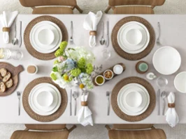Dining table set for six people with porcelain tableware from Villeroy & Boch, brown placemats, white napkins and a flower arrangement. Bread, spreads and condiments are included.