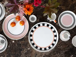 Service de table Memphis de Villeroy & Boch avec des assiettes décoratives, une tasse, des tasses et des prunes sur une surface en marbre brun décorée de fleurs brillantes.
