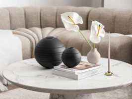 Die schwarze La Boule von Villeroy & Boch, einer beigen Vase mit weißen Blumen, einem Kerzenständer und einigen Büchern in einem beigen Wohnzimmerambiente.