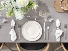 La table est dressée avec des assiettes blanches, des serviettes, des couverts, des verres et une composition florale sur une nappe grise.
