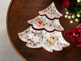 Overhead view of a 3-piece Christmas tree-shaped porcelain snack bowl set from Toy's Delight collection on a dark wooden table, filled with festive star cookies and a heart-shaped biscuit, next to a red gift box with a green-and-white striped ribbon and deep red flowers