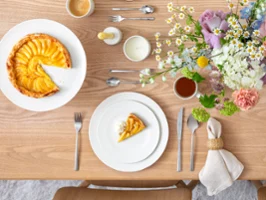 Ein gedeckter Tisch mit einem Stück Obsttorte auf einem For Me Teller Ein gedeckter Tisch mit einem Stück Obsttorte auf einem For Me Teller, einer ganzen Torte, Blumen und verschiedenen Getränken und Utensilien.