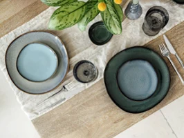 Vue de dessus d’une table moderne avec assiettes à dîner et à pain des collections Lave Vert, Lave Beige et Lave Glacé de Villeroy & Boch. La composition met en valeur les émaux réactifs uniques et les textures naturelles de chaque assiette sur une table neutre, sans nourriture, soulignant l’élégance artisanale du grès.