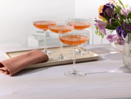 Vier Rose Garden Coupe-Gläser mit orangefarbenen Cocktails auf einem Tablett, daneben eine gefaltete Serviette und eine Vase mit Blumen auf einem weißen Tisch.