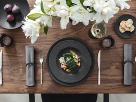 Mise en place élégante avec des assiettes Manufacture noires, des couverts, des figues, un plat de légumes et de crevettes, des serviettes grises, des fleurs blanches, un verre à vin et du pain coupé.
