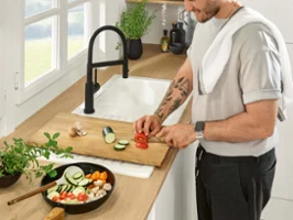 A man cuts vegetables on a wooden chopping board in a bright, modern kitchen. Various ingredients are laid out on the work surface and in a pan next to them.