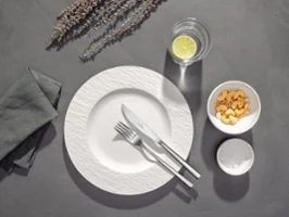 A white plate with fork and knife, a glass of water with lemon, a bowl of crackers and a napkin on a grey table.