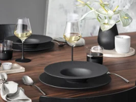 A modern wooden dining table set with Manufacture tableware, wine glasses, cutlery, and a vase with white flowers.