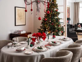 Festively decorated dining room with a table set for eight people. The table is set with festive crockery, candles and red flowers. Next to it is a Christmas tree with ornaments.