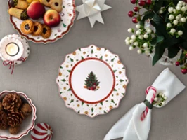 Toys Delight Plate A festively set table with a decorated plate featuring a Christmas tree motif, cookies, fruit, candles, napkins, and a flower arrangement.