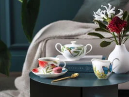 Elegant Amazonia tea service with colourful floral patterns on a round table, accompanied by a golden spoon, a white vase with flowers and stacked books in a cosy room ambience.