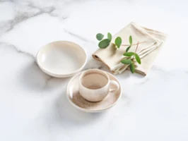 The Perlemor Sand espresso set, featuring a cup, saucer, and small bowl, is paired with a sand linen napkin on a sleek marble table.