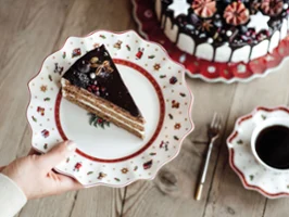 On the table are a decorated whole cake, a cup of coffee, a fork, and a saucer and plate set from Toy’s Delight. One hand holds a plate with a piece of chocolate cake. The table also features a decorated whole cake, a cup of coffee, a fork and a saucer, and plate set from Toy’s Delight.