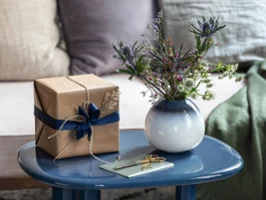 A white Lave vase stands on a blue table On a blue table is a gift wrapped in brown paper with a blue bow, a white lavender vase with flowers and a card.