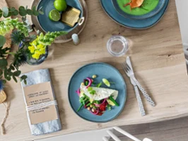 A blue plate with fresh salad, cutlery, a napkin and decorative tableware on a wooden table.