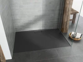 Grey Architectura shower tray with grey floor and wall tiles A modern walk-in shower with a black floor, grey tiled walls, a glass partition wall and a wooden beam. In the corner is a wooden stool with a basket of towels and soap.