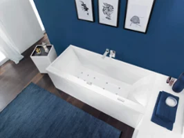 Une salle de bains moderne avec une baignoire rectangulaire blanche, des murs bleu foncé ornés d’œuvres d’art encadrées, un tapis bleu foncé, une table d’appoint avec des objets décoratifs et des serviettes de bain pliées sur une étagère blanche.
