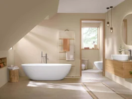 Un intérieur de salle de bains moderne avec une baignoire en îlot, un meuble en bois avec une vasque à poser, des toilettes, des miroirs ronds et des murs beige, avec de la lumière naturelle qui entre par une porte ouverte.