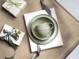 A green Perlemor cup, bowl and plate on gifts wrapped with grey ribbons, arranged on a beige textured surface.