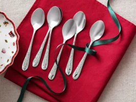 Six silver spoons are arranged on a red napkin, with a green bow wrapped around two of the spoons. On the left, part of a decorative Villeroy & Boch Toy's Delight bowl with Christmas patterns is visible.