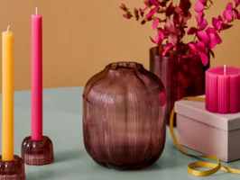 Nahaufnahme von geriffelten Glasvasen in Beerentönen, mit bunten Stabkerzen in passenden Glashaltern und einer rosa Stumpenkerze auf einer Geschenkbox.